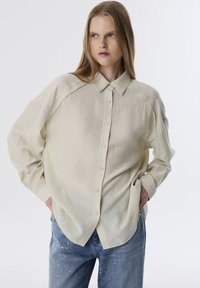 Lichtbeige knoopshirt in een gestructureerde stof met een iets oversized pasvorm, voorzien van een puntkraag en subtiele zilveren stippenversieringen.