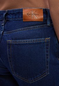 Tumši zili džinsi ir ar brūnu ādas jostas etiķeti, kurā ir iepresēts "Calvin Klein". Redzami šuvumi un aizmugures kabatas detaļas.