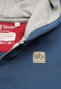 Blå og grå hættetrøje med lynlås, har en blød tekstur, rød inderside og et beige stoflabel med "B R" logo broderet på brystet.