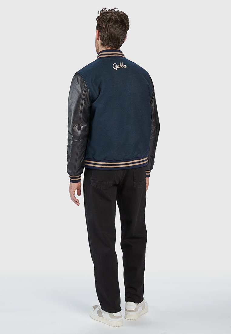 Marineblauwe wolmix varsityjack met zwarte leren mouwen, ribgebreide manchetten, gestreepte onderkant en "Gabba" geborduurd op de achterkant.