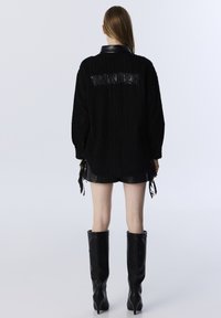 Zwarte gestructureerde oversized shirt met een leren kraag, voorzien van 'WONDER' in reliëf op de achterkant, gecombineerd met zwarte leren shorts en hoge laarzen.