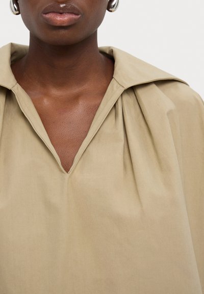 Blusa color tan con scollo a V, ampia colletto e dettagli in tessuto arricciato. La superficie liscia sottolinea la semplicità e l'eleganza.