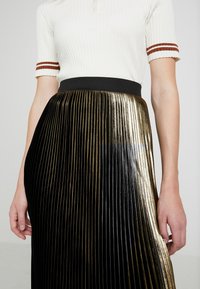 Gonna plissettata nera e oro con una texture lucida e una cintura nera in vita. La parte superiore presenta un design a coste bianco con strisce marroni sulle maniche.