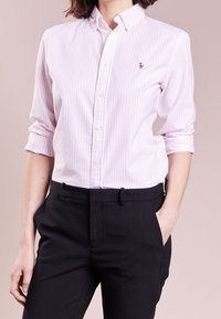 Ljusrosa randig skjorta med button-down krage, försedd med en liten broderad logotyp. Bärs tillsammans med svarta skräddarsydda byxor som visar en slät textur.