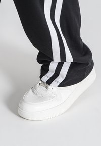 Hvide sneakers med krokodilletekstur, præget med sølvhardware og en tekstureret sål, parret med sorte bukser med hvide striber.