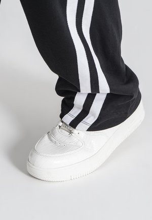 Hvide sneakers med krokodilletekstur, præget med sølvhardware og en tekstureret sål, parret med sorte bukser med hvide striber.