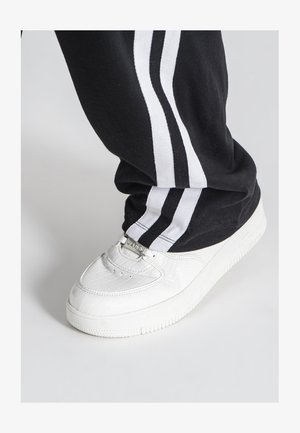 Witte sneakers met een krokodillenstructuur, voorzien van zilverkleurige hardware en een gestructureerde zool, gecombineerd met zwarte broek met witte strepen.