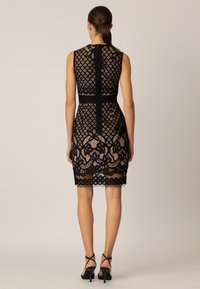 Robe en dentelle noire avec motif géométrique en losange et bordure en dentelle florale. Coupe ajustée, sans manches, longueur au genou, avec une fermeture éclair au dos.