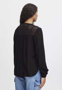 Svart blus med en texturerad, honungsbi-lace bakdel, långa ärmar med elastiska manschetter och en rundad nederkant. Slätt tyg med en avslappnad passform.