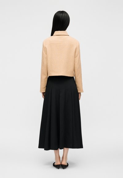 Femme aux longs cheveux noirs portant une veste courte beige et une jupe midi plissée noire, debout de dos sur un fond gris uni.