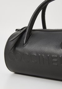 Sac à main en cuir noir avec un design arrondi, une surface texturée et un logo en relief. Il dispose de deux poignées courtes et d'une fermeture éclair.