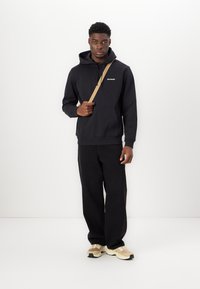 Sweatshirt à capuche noire avec poche avant et logo. Associé à un pantalon noir ample et des baskets beiges. Design simple et décontracté.