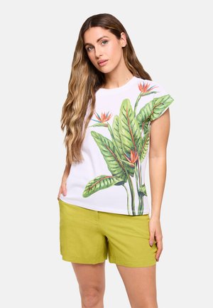 Mujer con el pelo largo y ondulado que lleva una camiseta blanca de manga corta con estampado de hojas tropicales verdes y naranjas y pantalones cortos verde claro, de pie con la mano en el bolsillo.