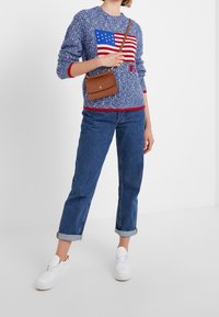 Pull en tricot bleu avec un motif de drapeau, accents rouges et coupe décontractée ; associé à un jean en denim avec les poignets retroussés et des baskets blanches. Sac à main marron.