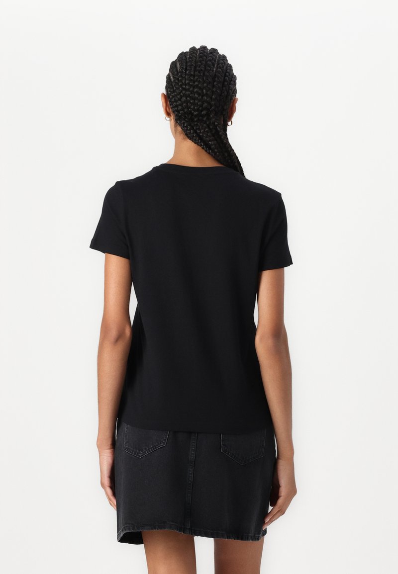 Schwarzes T-Shirt mit kurzen Ärmeln und rundem Ausschnitt, aus weicher Baumwolle gefertigt. Das Modell trägt es mit einem Jeansrock, der über Gesäßtaschen verfügt.