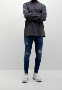 Šedá košile s dlouhým rukávem, potrhané modré skinny džíny a bílé tenisky s modrými detaily. Neformální outfit s uvolněným střihem.