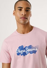 Svetle růžové bavlněné tričko s modro-bílým stříkacím designem a nápisem "Pepe Jeans LONDON" na přední straně. Krátké rukávy.