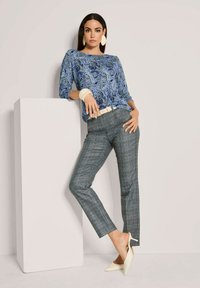 Frau trägt eine blaue Paisley-Bluse, graue Karohose, einen weißen Gürtel, beigefarbene High Heels, große Ohrringe und ein Armband, während sie sich an ein weißes Podest lehnt.
