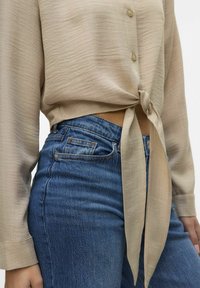 Camicia beige a maniche lunghe con nodo in vita, abbinata a jeans in denim blu a vita alta con tasche e leggere sfumature.
