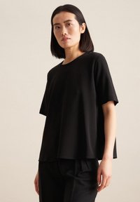 Top nero a maniche corte in una vestibilità rilassata, con scollo rotondo e tessuto leggero e morbido. Indossato con pantaloni neri.