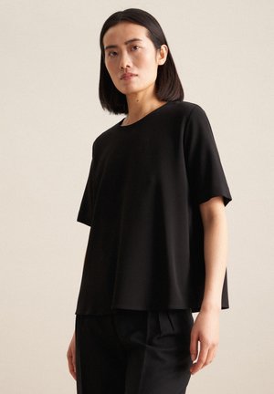 Top nero a maniche corte in una vestibilità rilassata, con scollo rotondo e tessuto leggero e morbido. Indossato con pantaloni neri.