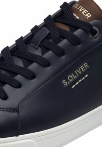 Baskets en cuir bleu marine avec semelle en caoutchouc blanc, bout rond et lacets noirs. Présente un logo doré et un accent marron au talon. Texture lisse.