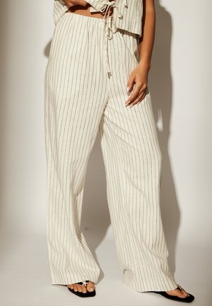 Pantaloni larghi in lino color crema con strisce verticali nere. Abbinati a un top corto coordinato, con dettagli a cravatta. Sandali neri piatti visibili.