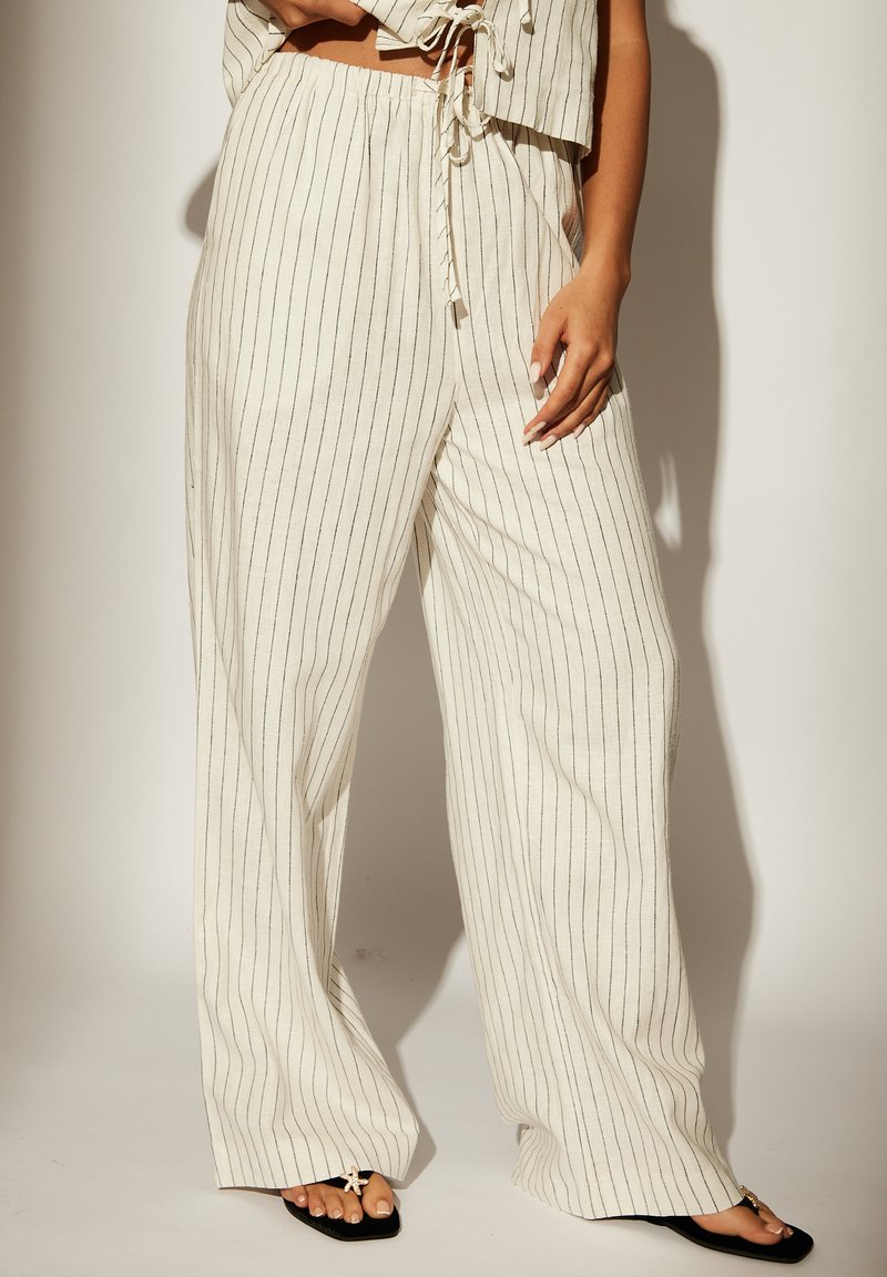 Pantalones de lino de pierna ancha en crema con rayas verticales negras. Combinados con un top corto a juego, que presenta un detalle de lazo. Sandalias planas negras visibles.