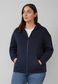 Marineblauwe zip-up hoodie gemaakt van zachte stof. Heeft een grote capuchon, voorzakken, geribde manchetten en een subtiele logodetail op de borst.