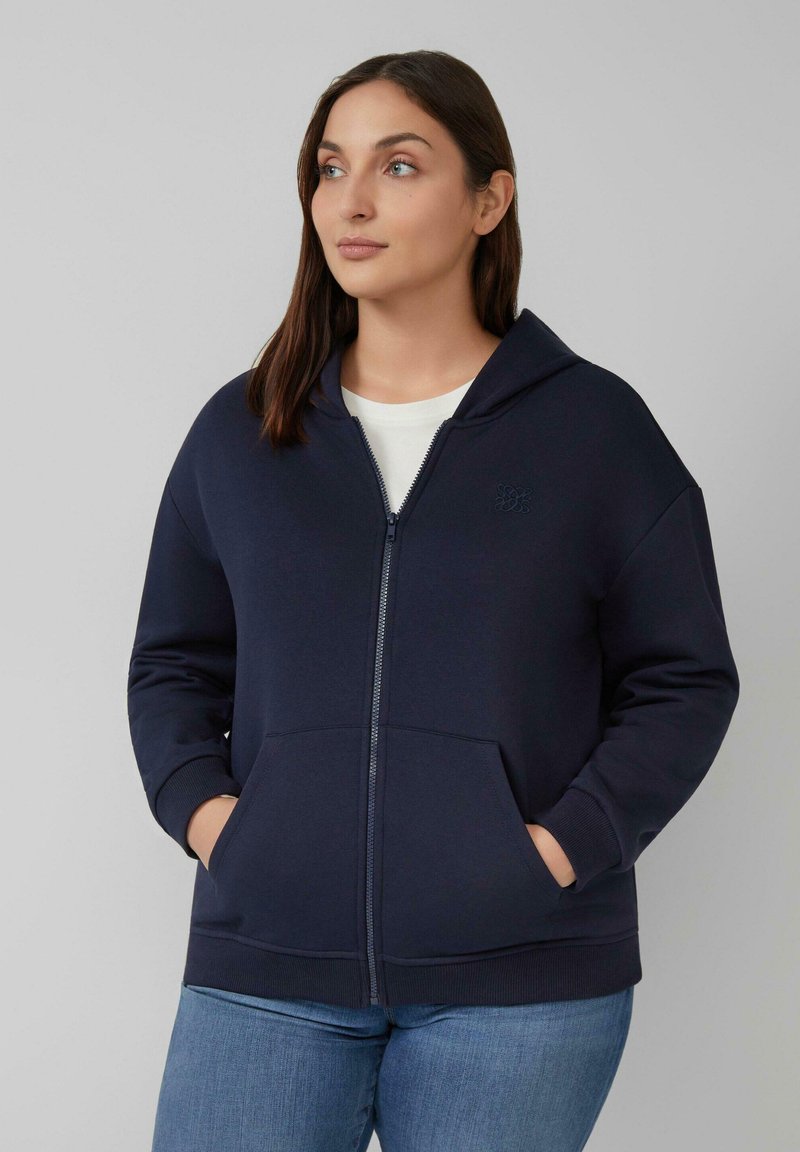 Marineblauwe zip-up hoodie gemaakt van zachte stof. Heeft een grote capuchon, voorzakken, geribde manchetten en een subtiele logodetail op de borst.