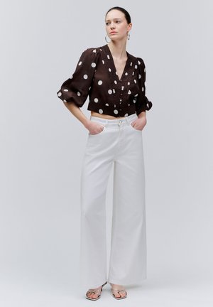 Žena v čiernej blúzke s bielymi bodkami a nafúknutými rukávmi, bielymi širokými nohavicami, striebornými sandálmi, ruky v vreckách, stojaca pred sivým pozadím.