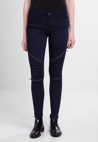 Námořnické skinny džíny s texturovaným designem, se šikmými zipovými akcenty na kolenou a falešnými kapsami, doplněné černými kotníkovými botami.