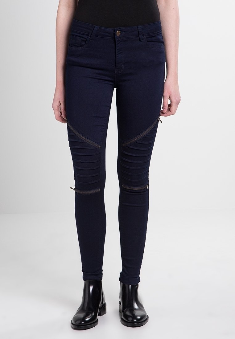 Námořnické skinny džíny s texturovaným designem, se šikmými zipovými akcenty na kolenou a falešnými kapsami, doplněné černými kotníkovými botami.