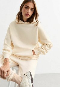 Krämfärgad hoodie tillverkad av mjukt tyg med en känguruficka, har oversized passform och ribbad mudd, i kombination med ljusa sneakers.