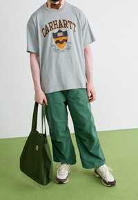 Graues Baumwoll-T-Shirt mit orangefarbenem "Carhartt"-Logo und Schild-Druck, kombiniert mit grünen lässigen Hosen und einer grünen Umhängetasche. Weiße Sneakers.
