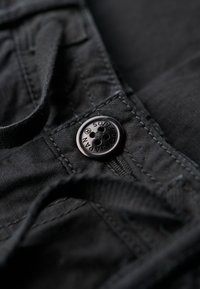 Zwart textieldetail met een ronde knoop met vier gaten, gestikt stof en zichtbare draadtextuur. Knoop met de tekst "Superdry."