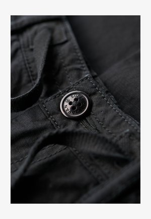 Zwarte stof met een close-up van een knoop met reliëfletters en vier gaten, gestikte details en een lichte textuur.