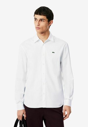 Joven con una camisa blanca abotonada con un pequeño logo verde de cocodrilo, sosteniendo la correa de una bolsa negra, mirando hacia su izquierda.
