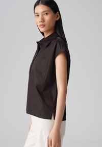 Dunkelbraunes Kurzarmhemd mit Kragen, ausgestattet mit einer Brusttasche und seitlichen Schlitzen. Glatte Textur. Kombiniert mit weißen Hosen.