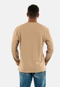 Camicia beige a maniche lunghe in tessuto morbido, con scollatura rotonda e vestibilità comoda, abbinata a jeans blu.