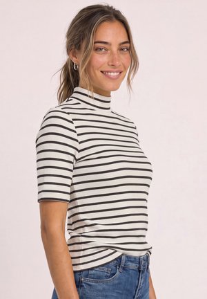 Ung kvinde smiler, iført en hvid og sort stribet kortærmet trøje og blå jeans, stående mod en ensfarvet lys baggrund.