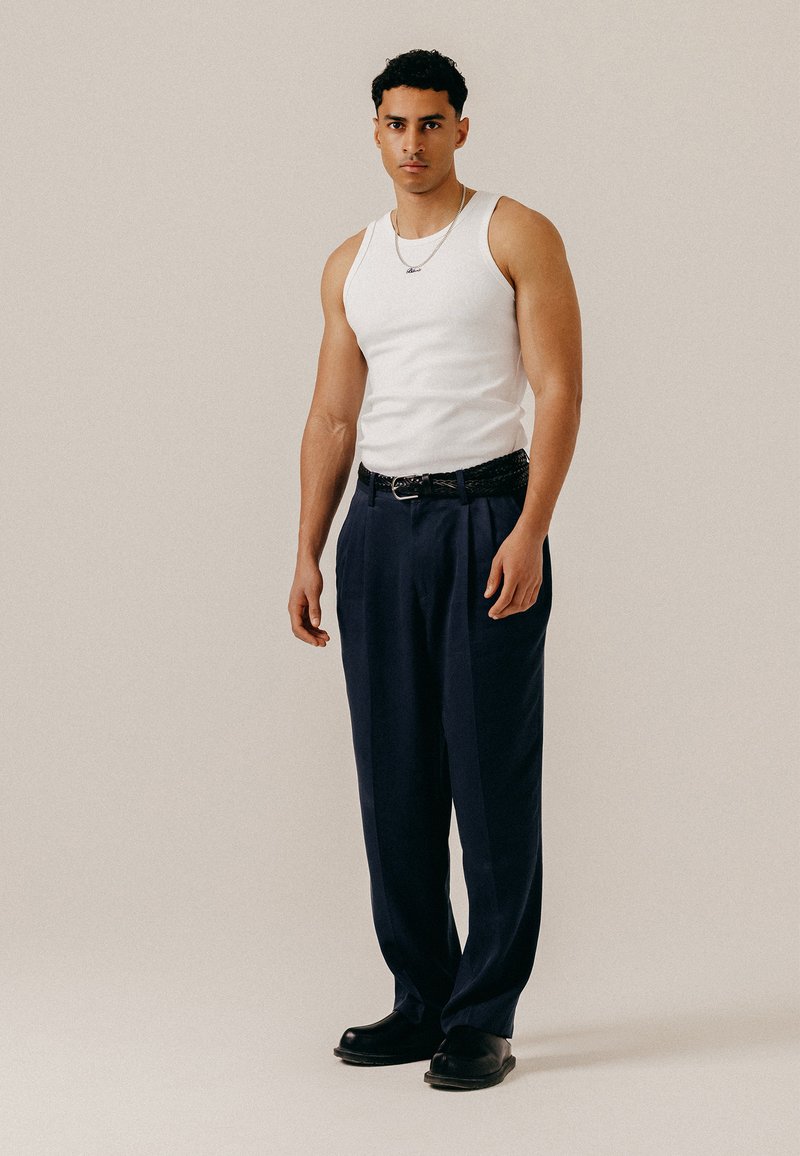 Jeune homme debout, portant un débardeur blanc sans manches, un pantalon plissé bleu marine avec une ceinture noire, des chaussures noires et un collier argenté.