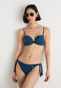 Teal bikini komplekts, kas ietver polsterētu krūšturi ar regulējamiem siksniņām un zelta akcentu aparatūru, apvienots ar sānu sasienamām biksītēm. Gluda, elastīga auduma.