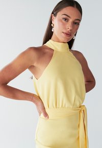 Kollane kaelapaeltega kleit, mis on vöökoha juures liibuva lõikega ja millel on lipsuaksent. Valmistatud pehmest kangast, mis on sileda tekstuuriga ja lihtsa disainiga.