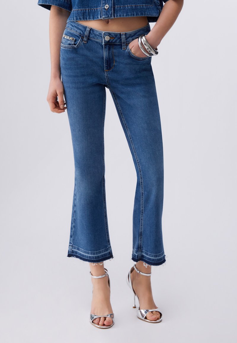 Femeie purtând blugi evazați scurtați din denim albastru, asortați cu sandale argintii cu toc înalt și brățări argintii la o încheietură.