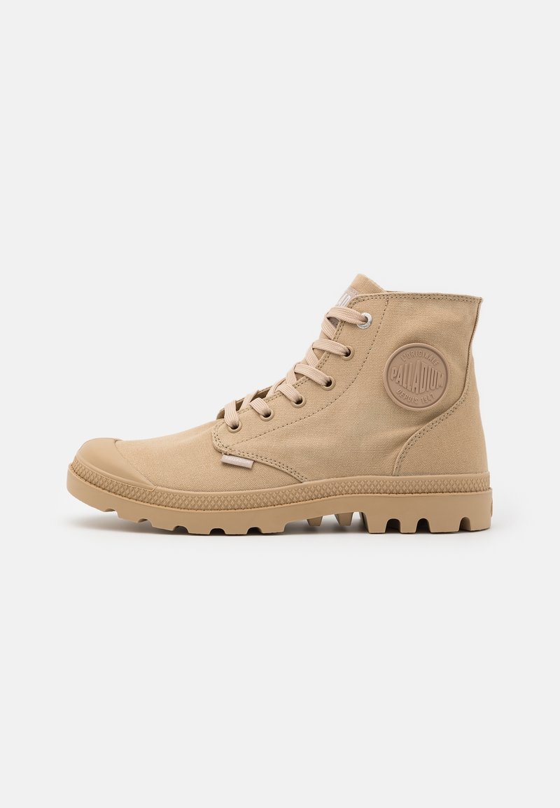 Botte montante beige en toile avec semelle en caoutchouc, fermeture à lacets à l'avant et écusson rond avec logo sur le côté de la cheville.