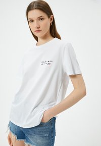 Hvid bomulds T-shirt med korte ærmer, prydet med et grafisk tryk, der inkluderer "CLEAN" og "SEA" samt røde accenttekster. Casual fit, rund halsudskæring.
