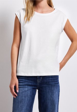 Woman wearing a plain white short-sleeve shirt and blue jeans, standing with one hand in pocket and the other relaxed by her side.