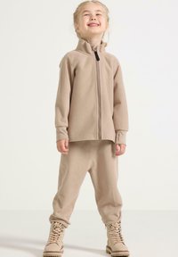 Completo in pile beige con giacca con zip e pantaloni abbinati. La giacca presenta un colletto alto, mentre i pantaloni hanno una vestibilità comoda. Stivali beige completano il look.