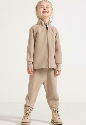 Completo in pile beige con giacca con zip e pantaloni abbinati. La giacca presenta un colletto alto, mentre i pantaloni hanno una vestibilità comoda. Stivali beige completano il look.